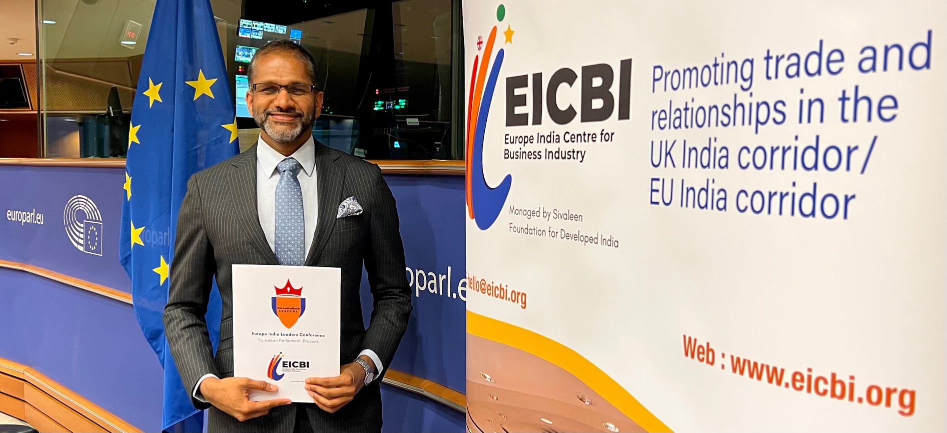 Rahul Venkit holding an EICBI folder in front of European Parliament flags, with the EICBI banner